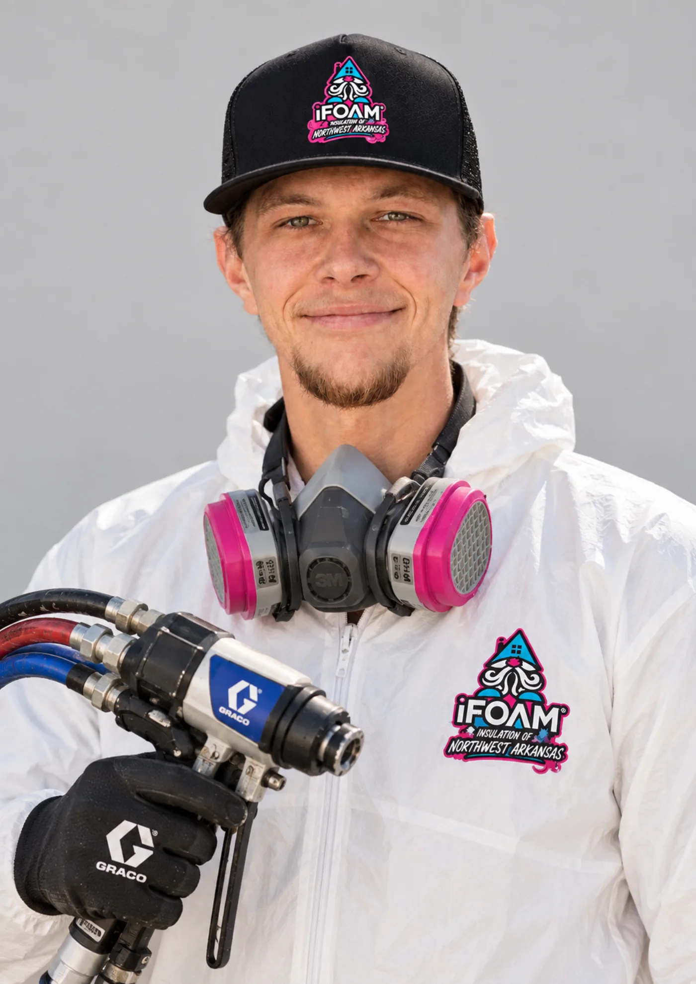 An iFOAM of Northwest Arkansas spray foam technician in a branded suit and cap with a half-mask respirator and spray gun
